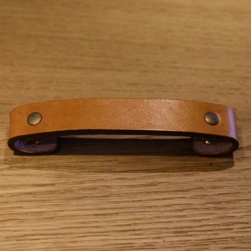 Brown leather drawer pulls with brass rivets on a wooden surface. Rustic cabinet handles.